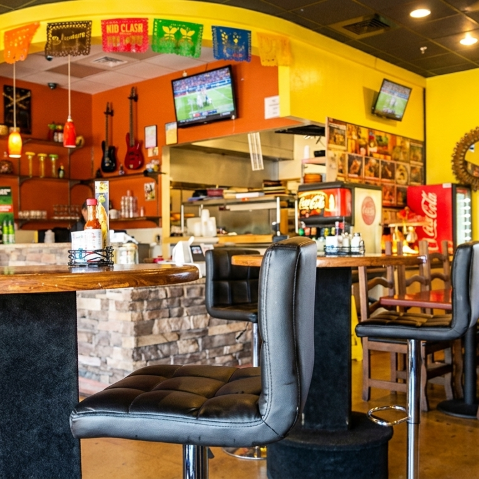 Colorful Mexican restaurant interior with bar seating, televisions, guitars, festive papel picado banners, and dining tables.